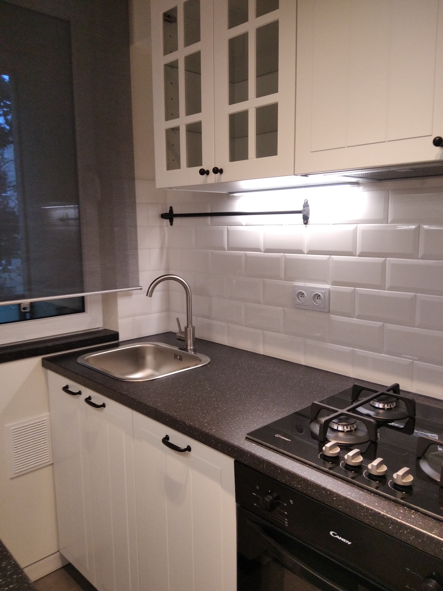 Fragment kuchni z białymi szafkami z przeszklonymi frontami, czarnym blatem, metalowym zlewem i płytą gazową, oświetlony paskiem LED pod szafkami.