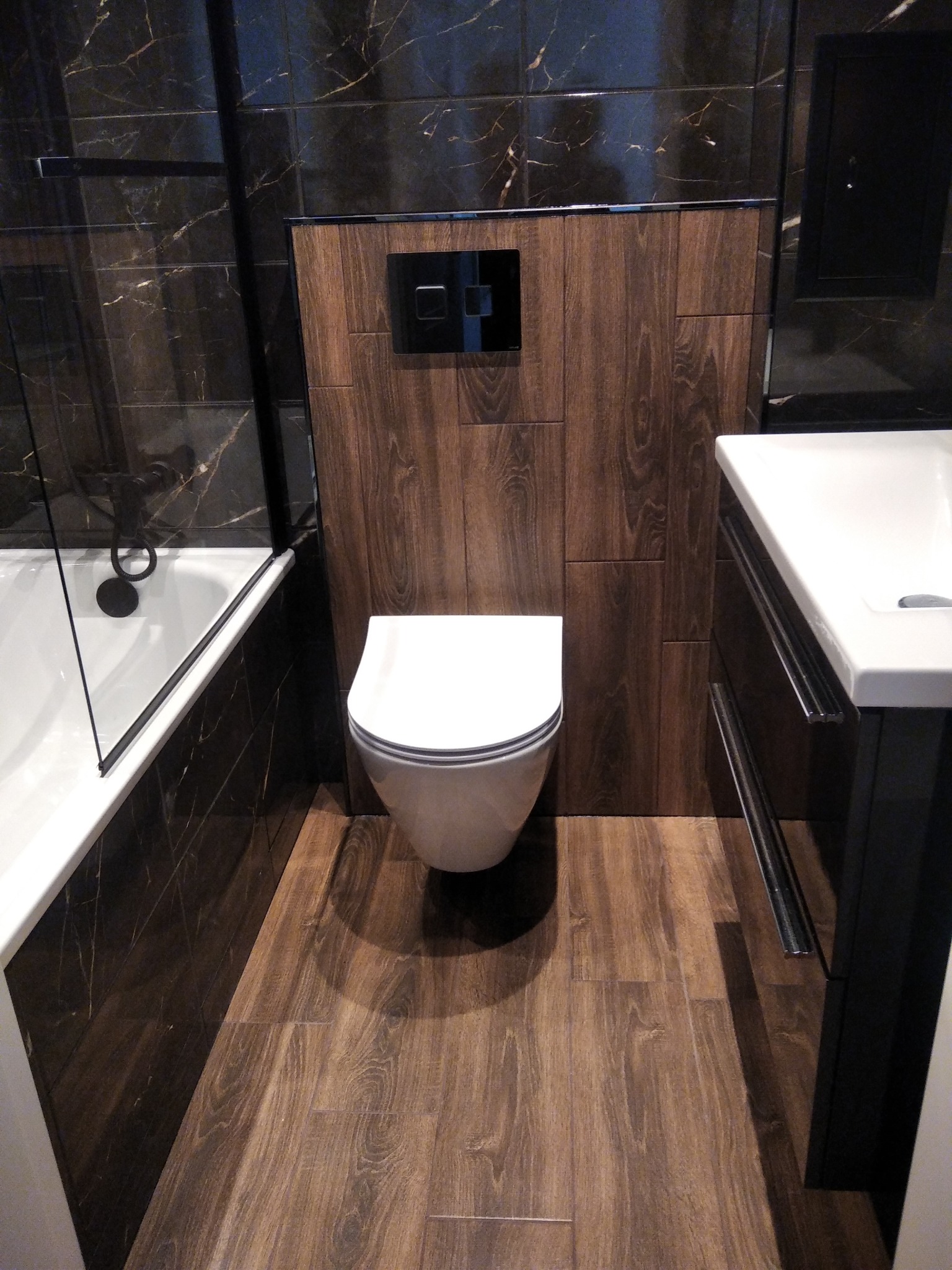 Nowoczesna łazienka z ciemnymi płytkami imitującymi marmur, drewnopodobną zabudową toalety i podłogą z paneli imitujących drewno, widoczna wanna z czarną armaturą i umywalka z szafką.