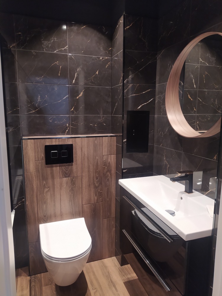 Nowoczesna łazienka z ciemnymi płytkami imitującymi marmur, okrągłym lustrem z drewnianą ramą, podwieszaną toaletą i szafką umywalkową z czarnym połyskiem.