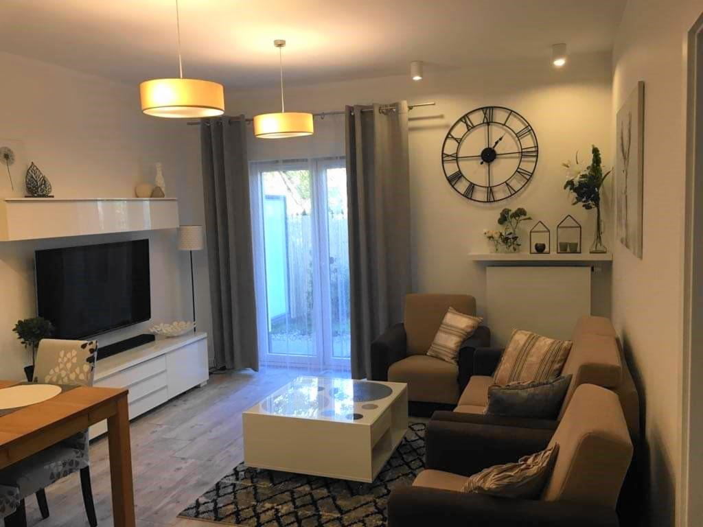 Salon w Warszawie po wykończeniu wnętrz, z drewnianym stołem w rogu, szarą sofą i fotelami, białymi meblami pod telewizorem i dużym zegarem na ścianie.