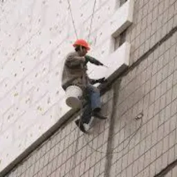 Pracownik w kasku, zawieszony na linach, mocuje białe płyty izolacyjne do ściany budynku z widoczną fakturą. W tle szara, pochmurna pogoda.