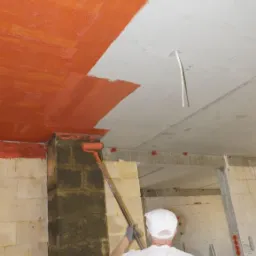 Pracownik w białej koszulce z napisem 'DEKOR-TYNK' maluje pomarańczową farbą sufit i filar z cegieł przy użyciu wałka na długim kiju w surowym wnętrzu budynku.