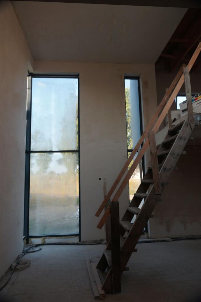 Wnętrze w trakcie prac wykończeniowych: ściany po nałożeniu tynku, widoczne duże okna i prowizoryczne drewniane schody.