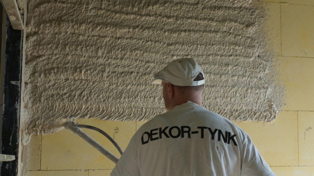 Aplikacja tynku maszynowego na ścianę z bloczków; widoczny fragment świeżo nałożonej warstwy z charakterystyczną fakturą oraz pracownik w czapce i koszulce z napisem DEKOR-TYNK.