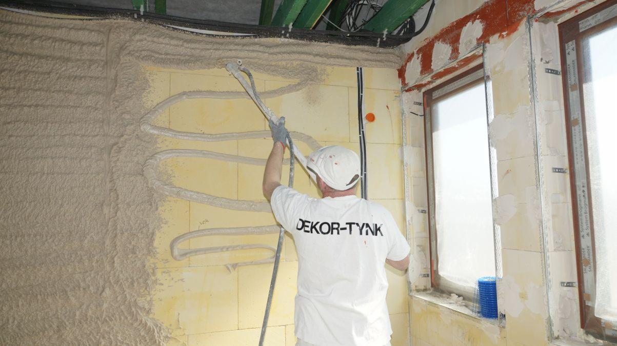 Tynkarz w białej koszulce z napisem DEKOR-TYNK nakłada tynk maszynowy na ścianę z instalacją grzewczą, widoczne okna z zabezpieczeniami.