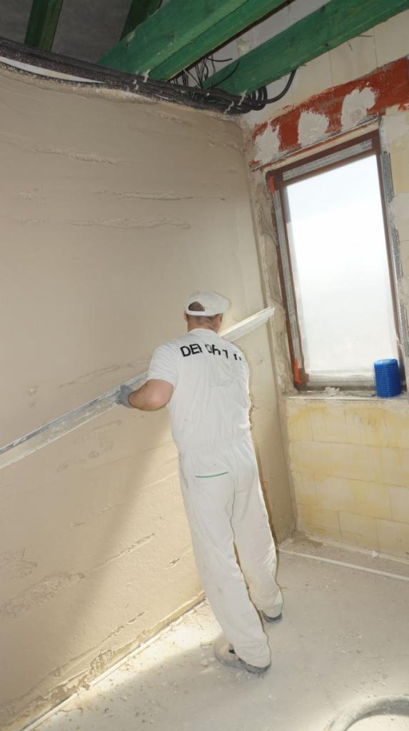 Pracownik w białym kombinezonie wygładza świeżo nałożony tynk na ścianie w pomieszczeniu z oknem, widoczne zielone elementy konstrukcji dachu i instalacja elektryczna pod sufitem.