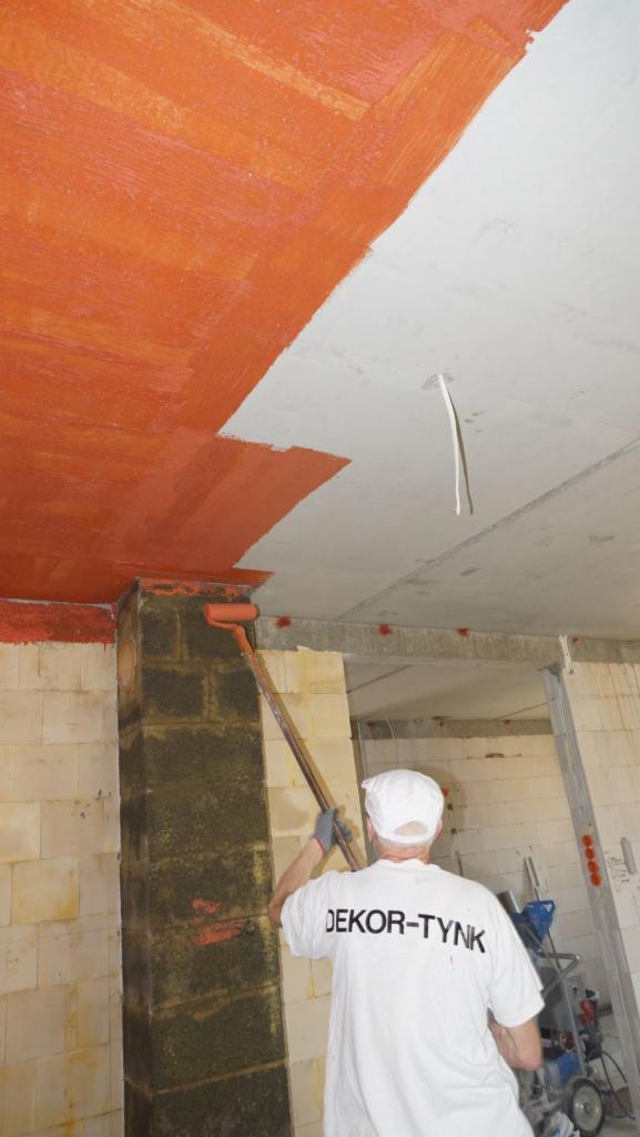 Pracownik w białej koszulce z napisem 'DEKOR-TYNK' maluje pomarańczową farbą sufit i filar z cegieł przy użyciu wałka na długim kiju w surowym wnętrzu budynku.