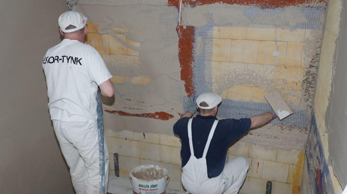 Dwaj pracownicy w strojach roboczych nanoszą zaprawę na ścianę z siatką wzmacniającą; widoczny częściowo otynkowany fragment ściany i wiadro z zaprawą.