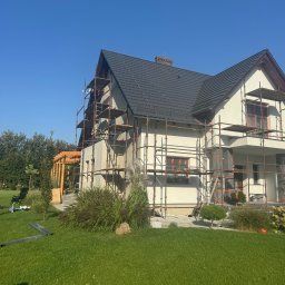 Bud-Mar Marcin Wlazik - Dwukondygnacyjny dom w trakcie prac elewacyjnych, otoczony rusztowaniami, z widoczną drewnianą pergolą w ogrodzie, w słoneczny dzień.