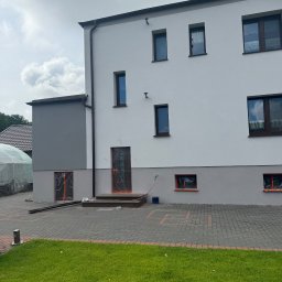 Bud-Mar Marcin Wlazik - Dwukondygnacyjny dom z jasną elewacją w trakcie prac wykończeniowych, okna i drzwi zabezpieczone folią, widoczny fragment ogrodu i kostki brukowej.