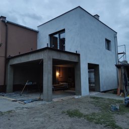 Bud-Mar Marcin Wlazik - Nowoczesny dom w trakcie prac elewacyjnych, widoczne świeżo nałożone tynki na ścianach i podbiciu garażu, teren wokół częściowo zabezpieczony folią, w tle widoczne rusztowanie.