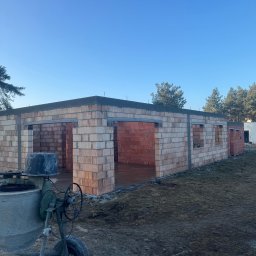 Bud-Mar Marcin Wlazik - Budowa domu z czerwonej cegły, widoczna betoniarka. Ściany w trakcie murowania, z otworami okiennymi. Teren budowy, las w tle, błękitne niebo.