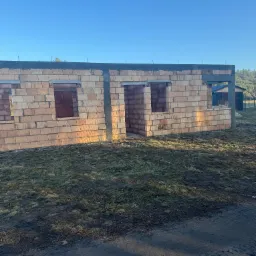 Wznoszenie murów z czerwonej cegły porotherm na budowie domu jednorodzinnego w słoneczny dzień. Widoczne otwory okienne i drzwiowe.