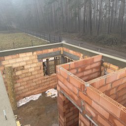 Bud-Mar Marcin Wlazik - Wznoszenie ścian domu z czerwonej cegły ceramicznej na fundamencie. Widok z góry na budowę w lesie, fragment pracownika w otworze okiennym.