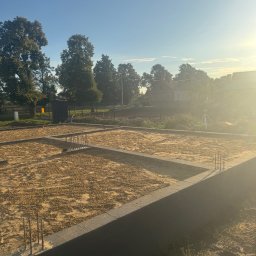 Bud-Mar Marcin Wlazik - Zalane fundamenty pod budowę domu jednorodzinnego, z widocznym zbrojeniem i piaskiem. W tle drzewa i zabudowania w promieniach wschodzącego słońca.