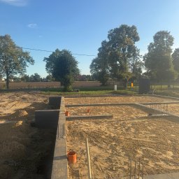 Bud-Mar Marcin Wlazik - Fundamenty domu w trakcie budowy, widoczne szalunki, zbrojenia i piasek. Elementy instalacji wystają z ziemi. W tle drzewa i błękitne niebo.