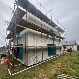 Bud-Mar Marcin Wlazik - Dwukondygnacyjny dom w trakcie ocieplania, otoczony rusztowaniem. Okna zabezpieczone folią. Na zewnątrz pracownicy budowlani i trawa.