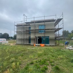 Bud-Mar Marcin Wlazik - Dwukondygnacyjny dom w trakcie ocieplania elewacji, z rusztowaniami i pracownikiem przygotowującym zaprawę obok budynku. Okna i drzwi zabezpieczone folią.