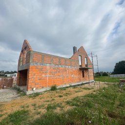 Bud-Mar Marcin Wlazik - Surowy stan domu z czerwonej cegły z kominem i rusztowaniem na tle pochmurnego nieba. Widoczne palety z cegłami i zielona trawa.