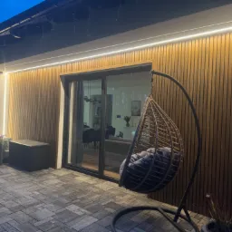 Nowoczesna elewacja domu z drewnianymi panelami, oświetleniem LED i szarą kostką brukową. Widoczny fragment tarasu z wiszącym fotelem i kwiatami.