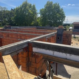 Bud-Mar Marcin Wlazik - Wznoszenie ścian z czerwonej cegły na budowie domu jednorodzinnego. Widoczne szalunki, narzędzia i elementy konstrukcyjne. W tle drzewa i turbina wiatrowa.