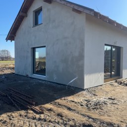 Bud-Mar Marcin Wlazik - Surowy, otynkowany dom jednorodzinny z drewnianą konstrukcją dachu, duże okna, teren wokół domu w trakcie prac ziemnych. Słoneczny dzień, błękitne niebo.