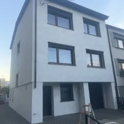 Ocieplony, dwukondygnacyjny dom z nową, jasną elewacją i brązowymi oknami. Na podjeździe stoi drewniana drabina. Widok z dołu, w tle niebieskie niebo.