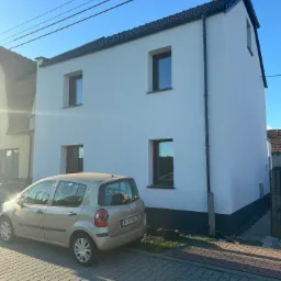 Biały dom z nową elewacją i ciemnymi oknami, zaparkowany samochód. Świeżo pomalowana fasada w słoneczny dzień. Widok na dom z chodnika.