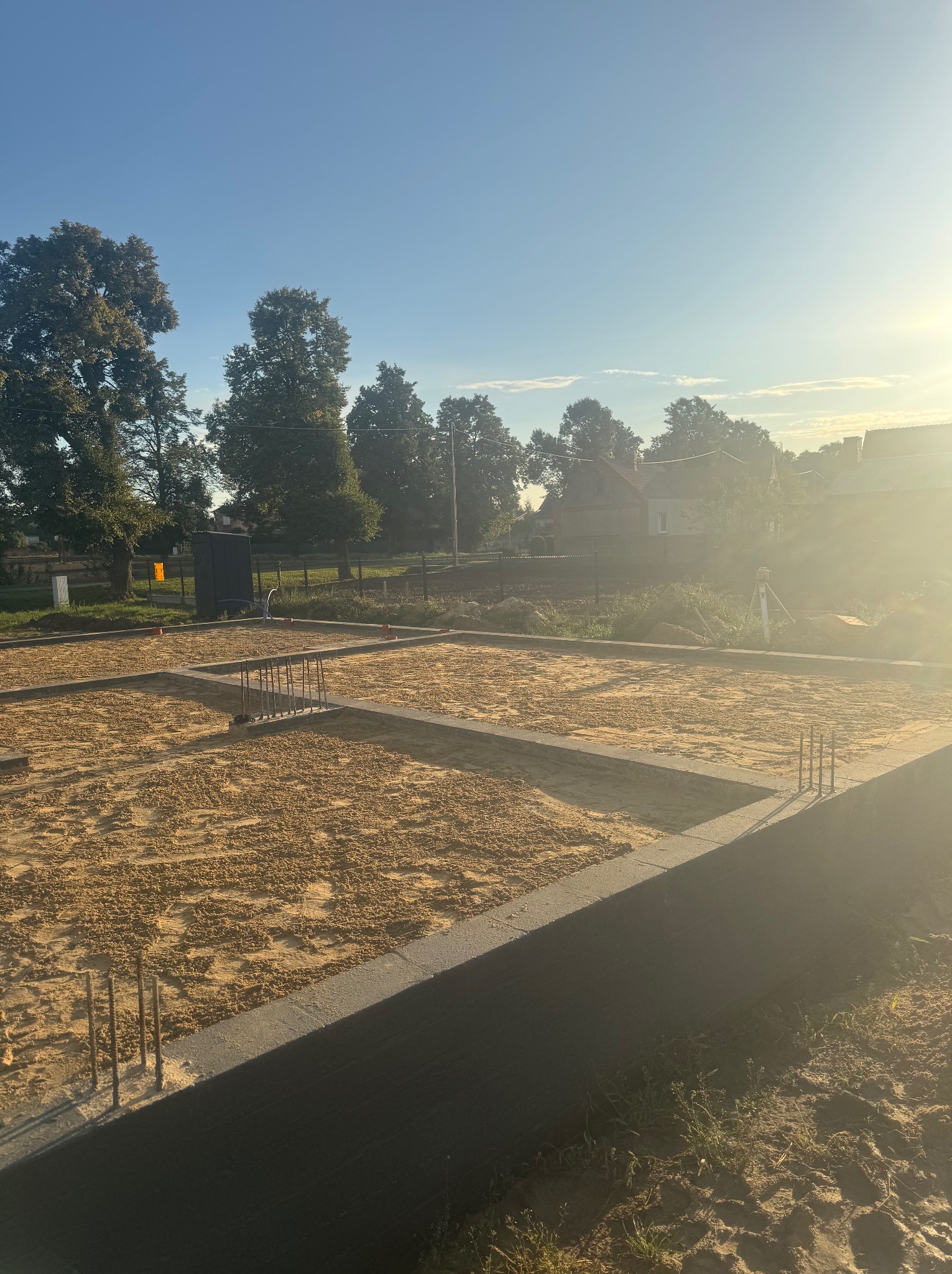 Zalane fundamenty pod budowę domu jednorodzinnego, z widocznym zbrojeniem i piaskiem. W tle drzewa i zabudowania w promieniach wschodzącego słońca.