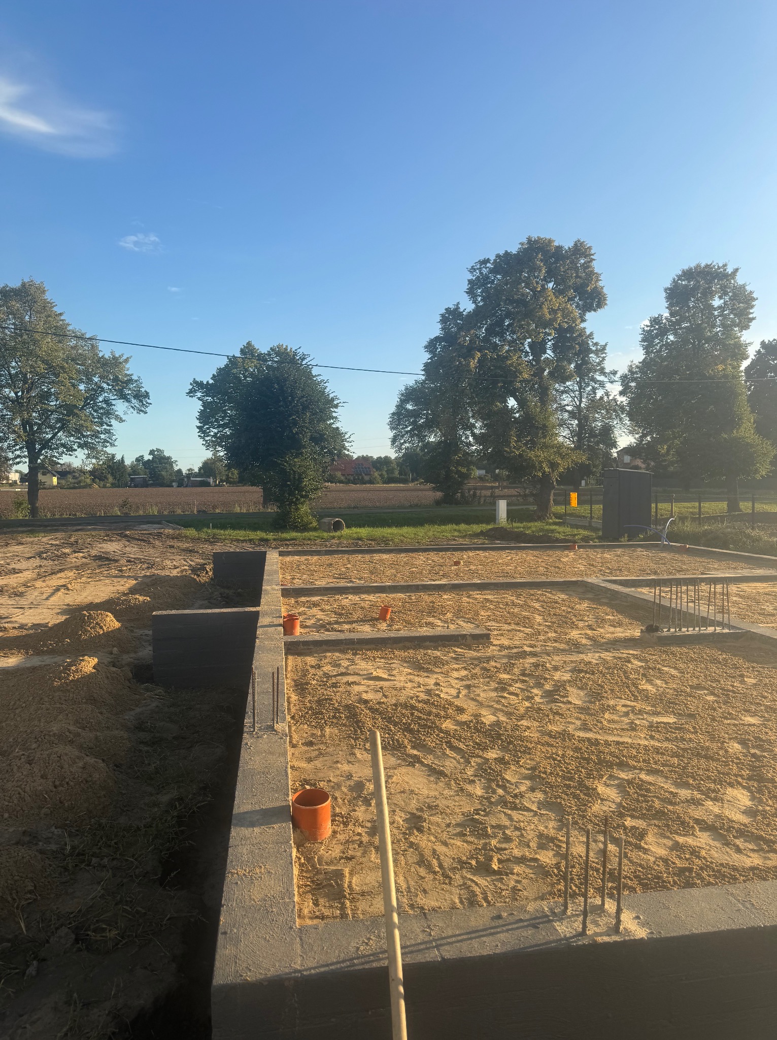 Fundamenty domu w trakcie budowy, widoczne szalunki, zbrojenia i piasek. Elementy instalacji wystają z ziemi. W tle drzewa i błękitne niebo.