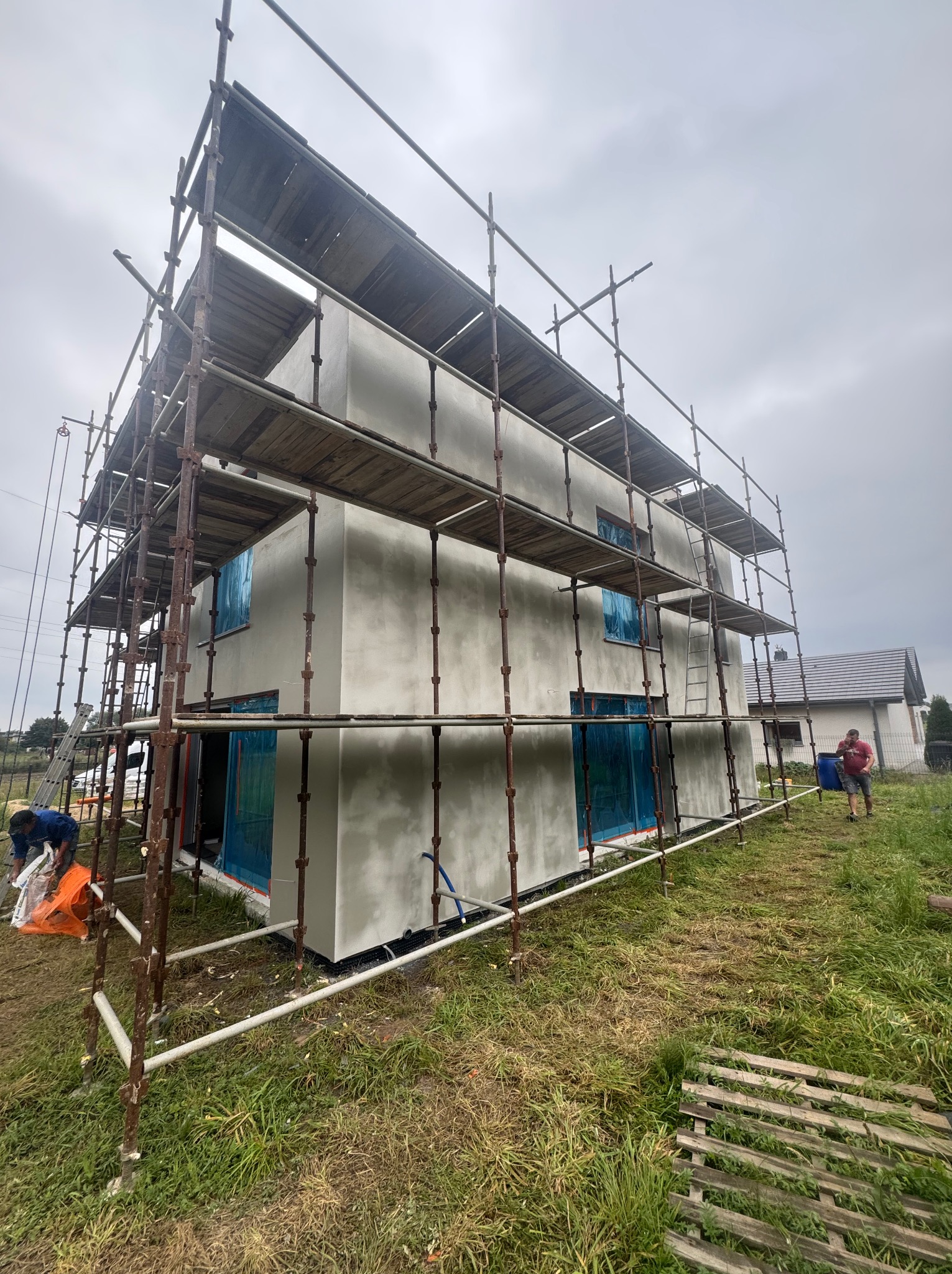 Dwukondygnacyjny dom w trakcie ocieplania, otoczony rusztowaniem. Okna zabezpieczone folią. Na zewnątrz pracownicy budowlani i trawa.