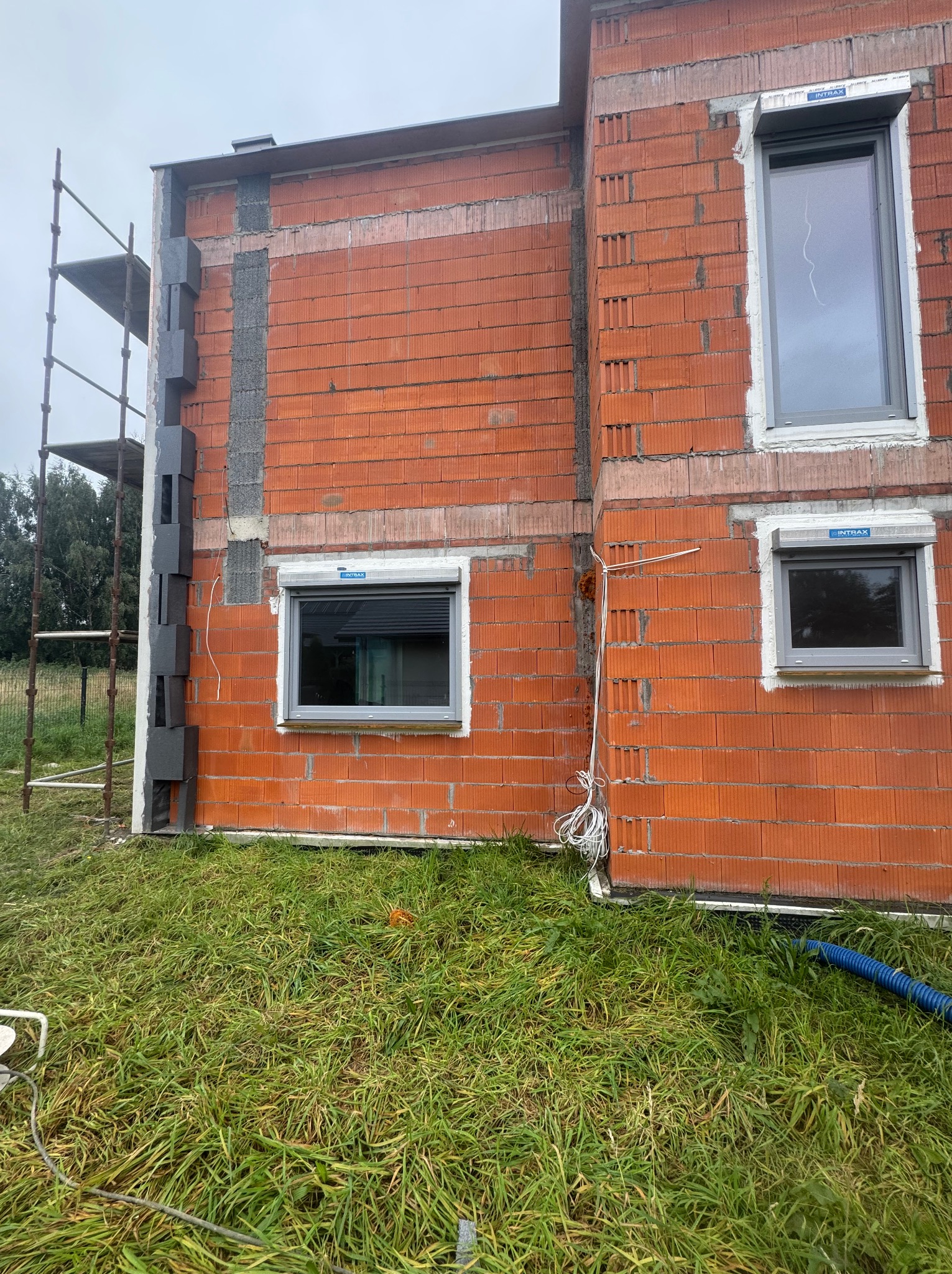 Surowy, czerwony mur z oknami z roletami, widoczne szare bloki izolacyjne na ścianie i rusztowanie. Budowa domu w trakcie, trawa przed budynkiem.