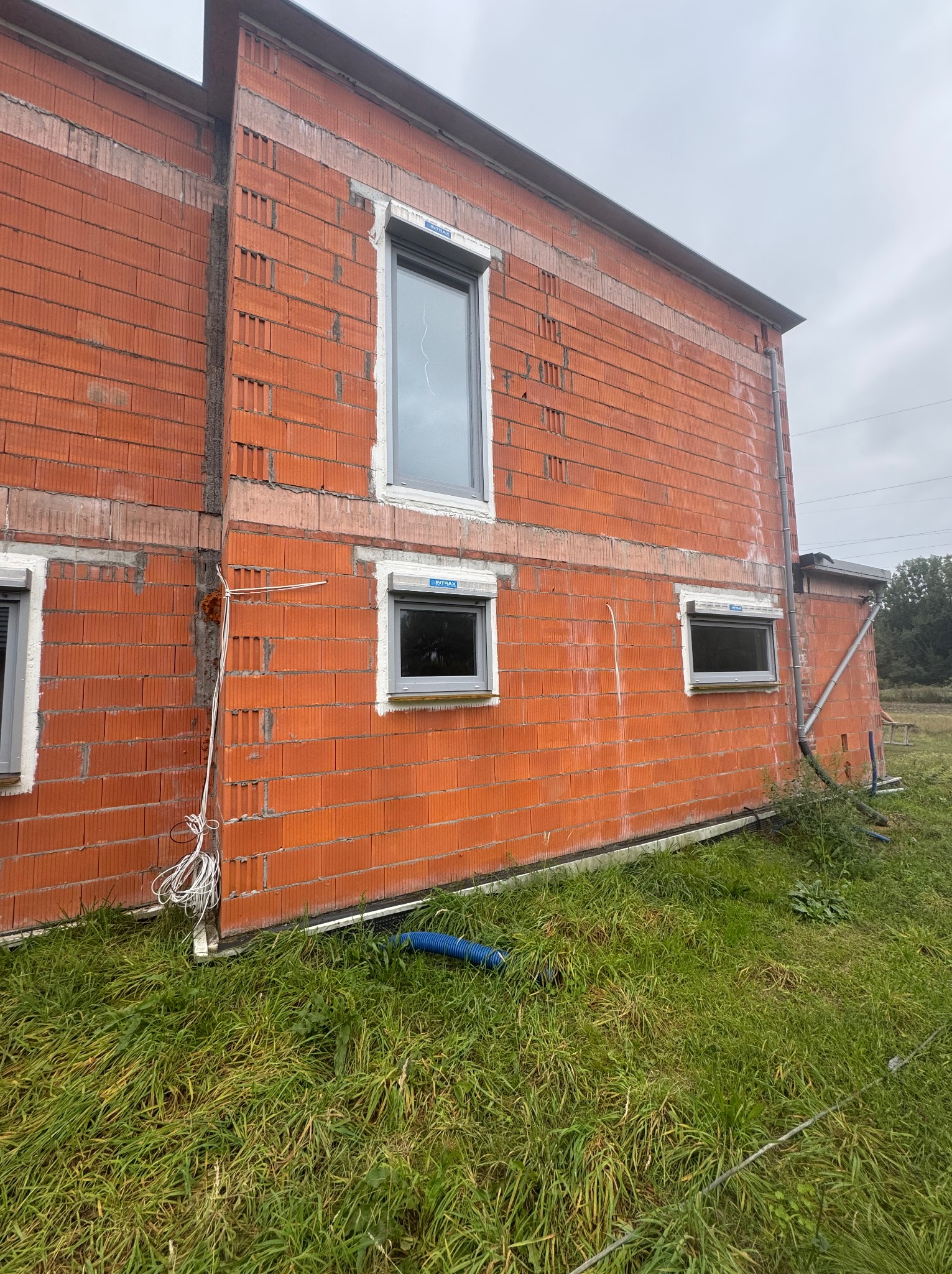 Surowy mur z pustaków ceramicznych z oknami w szarych ramach, widok z zewnątrz na tle zieleni. Częściowo otynkowane nadproża okienne. Budowa domu w toku.