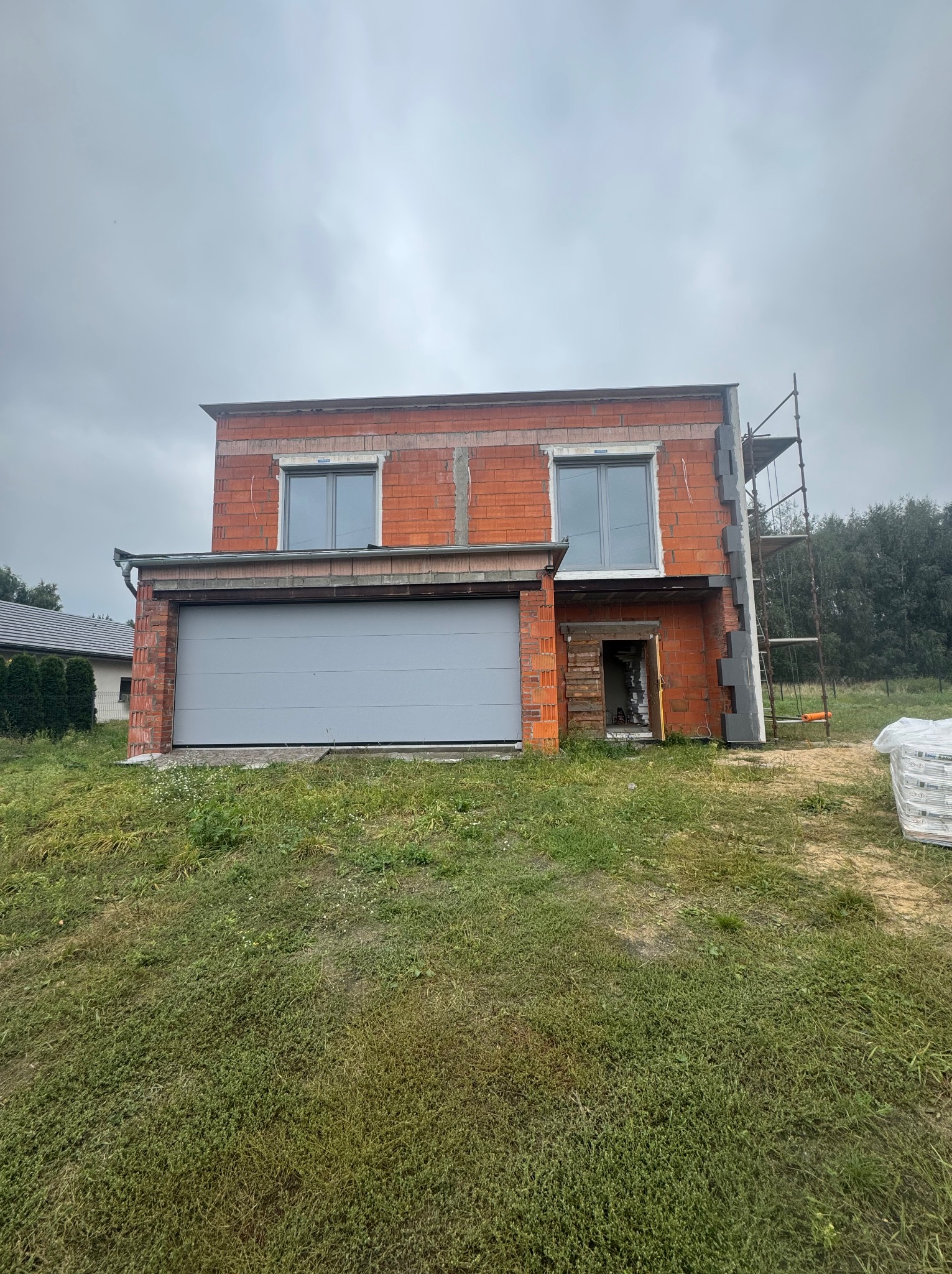 Dwukondygnacyjny dom w trakcie budowy z garażem, widoczne rusztowania i materiały izolacyjne. Częściowo ocieplona ściana. Okna zamontowane. Pochmurne niebo w tle.