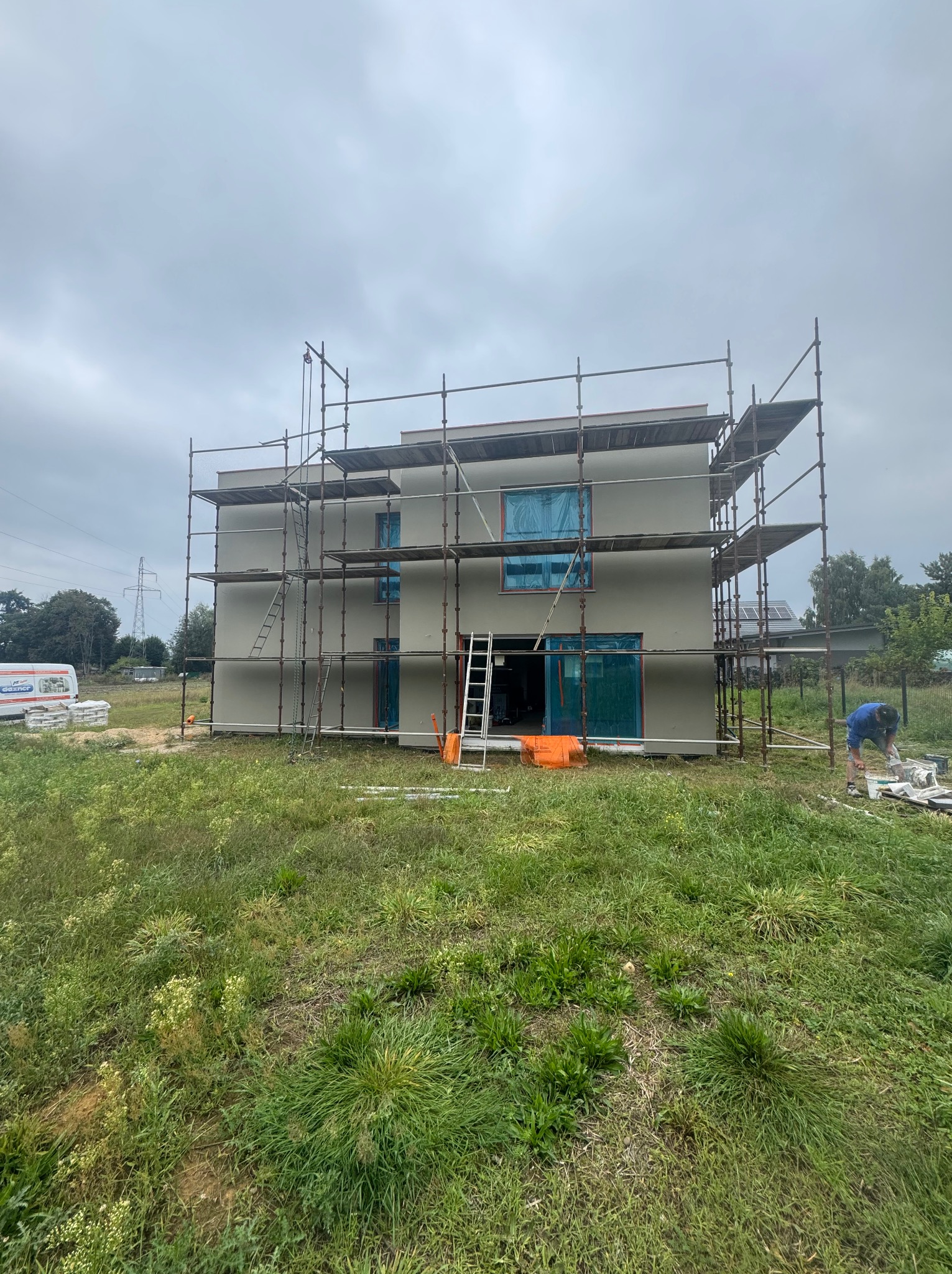 Dwukondygnacyjny dom w trakcie ocieplania elewacji, z rusztowaniami i pracownikiem przygotowującym zaprawę obok budynku. Okna i drzwi zabezpieczone folią.