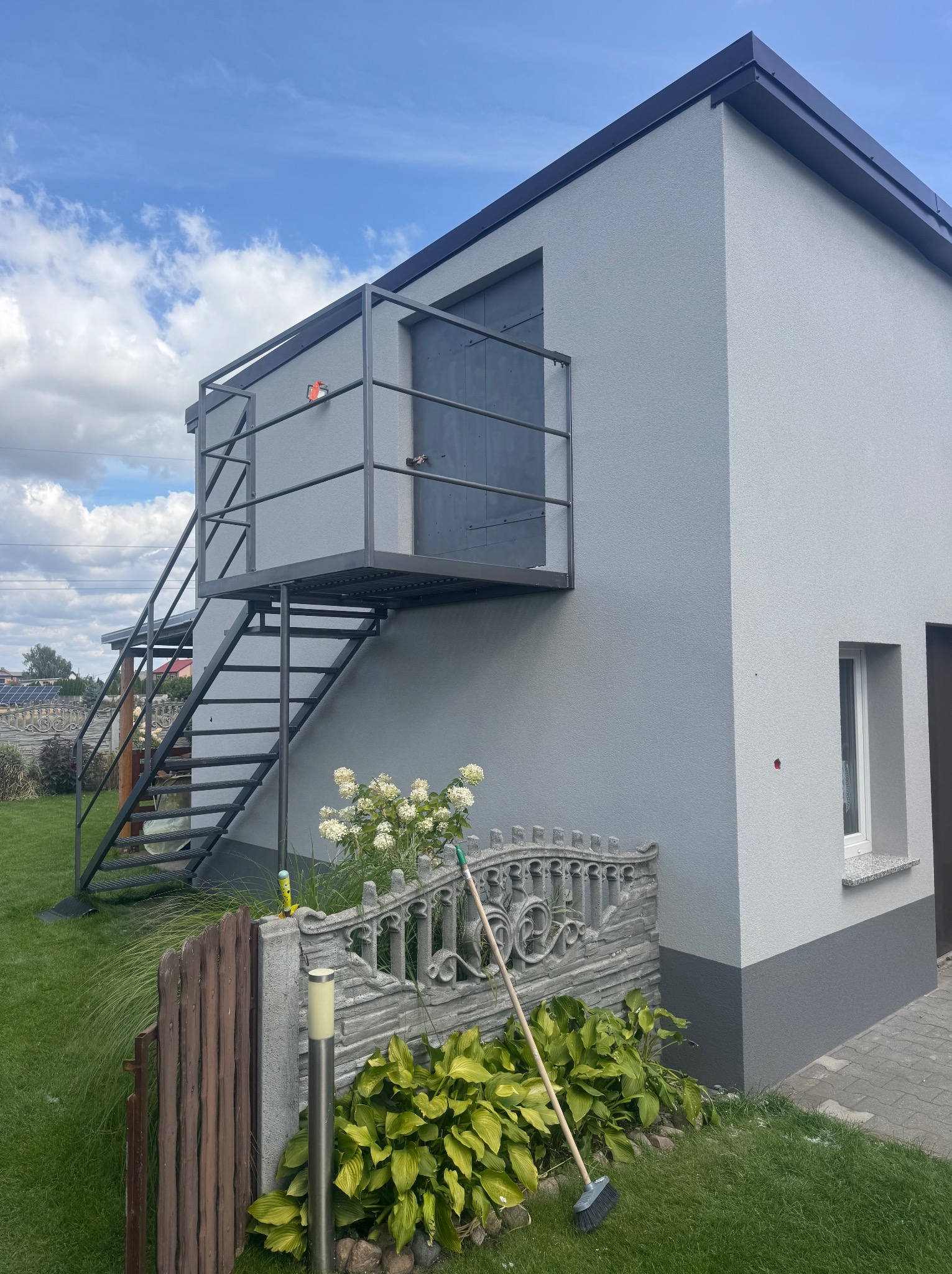 Szary budynek z metalowymi schodami zewnętrznymi i balkonem, widok z poziomu ogrodu. Świeżo pomalowana elewacja z widoczną fakturą tynku.