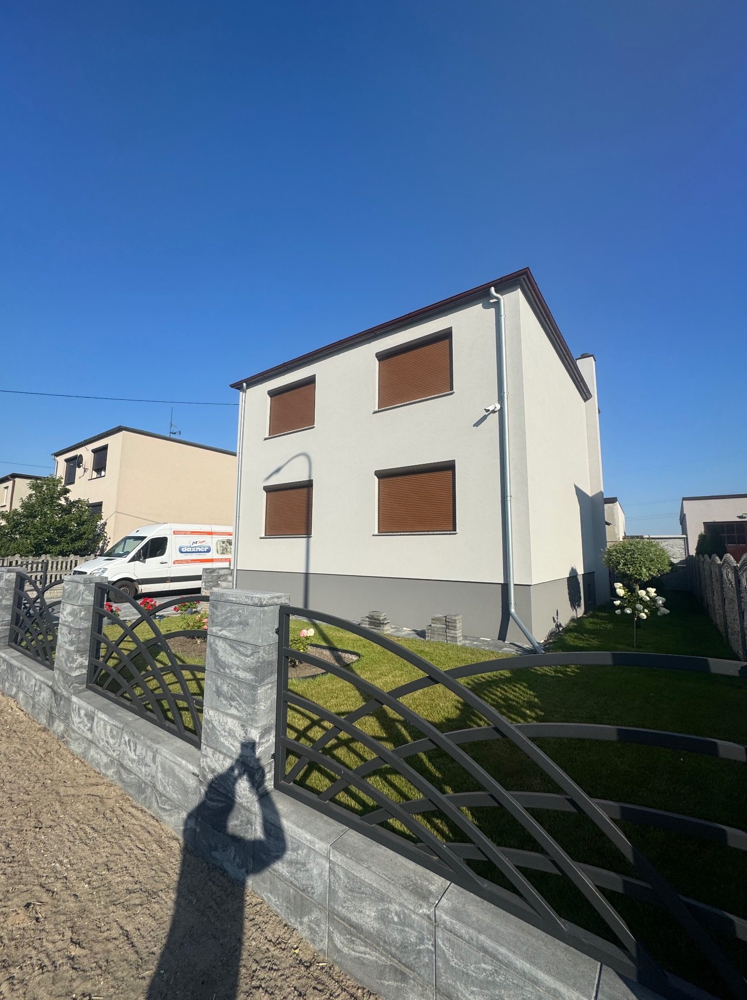 Dwukondygnacyjny dom z jasną elewacją i brązowymi roletami, widziany zza ozdobnego, metalowego ogrodzenia. W tle zaparkowany biały bus.