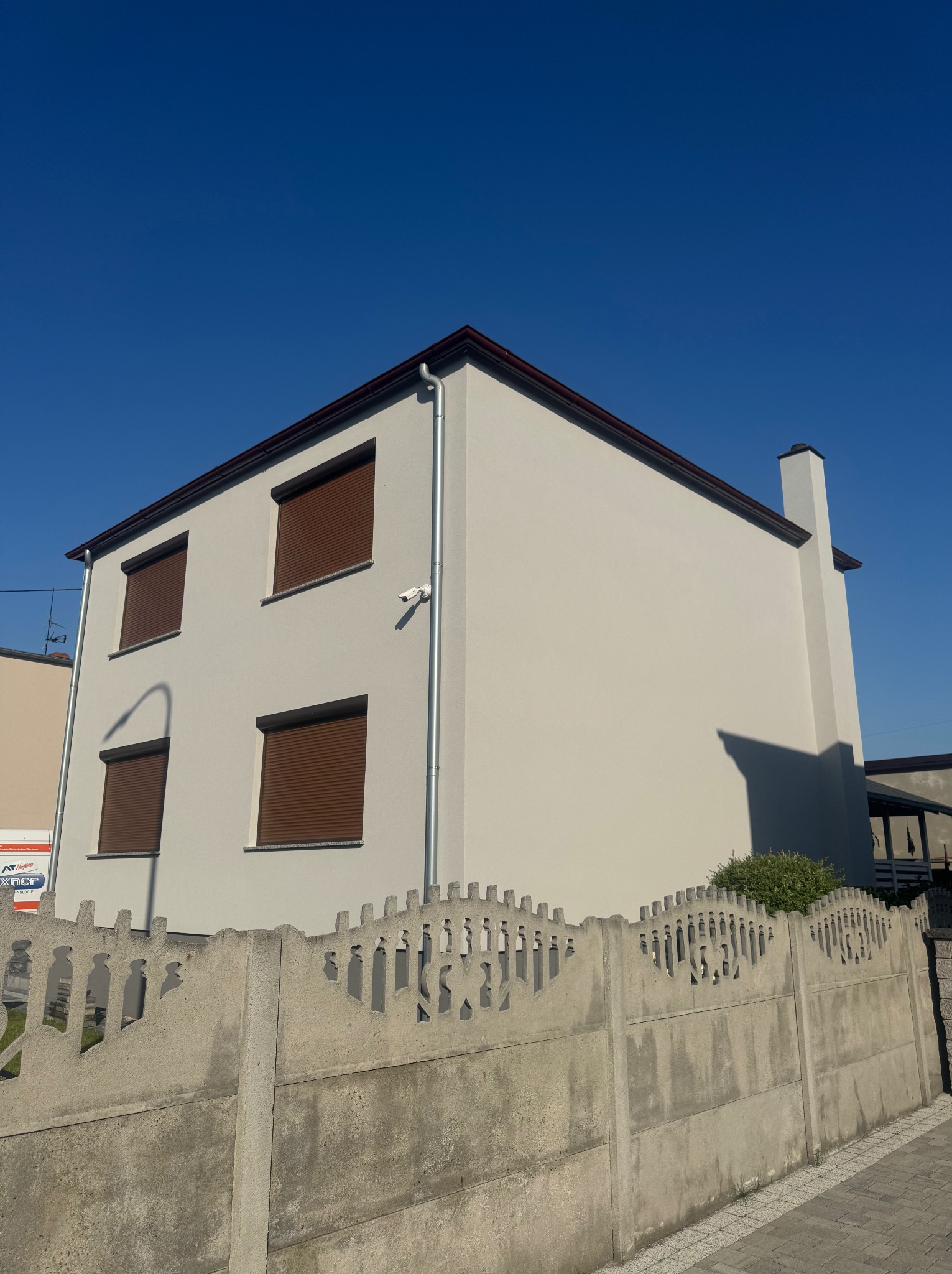 Dwupiętrowy dom z jasną elewacją i brązowymi roletami, widziany zza betonowego ogrodzenia o dekoracyjnym wzorze. Czyste, błękitne niebo w tle.
