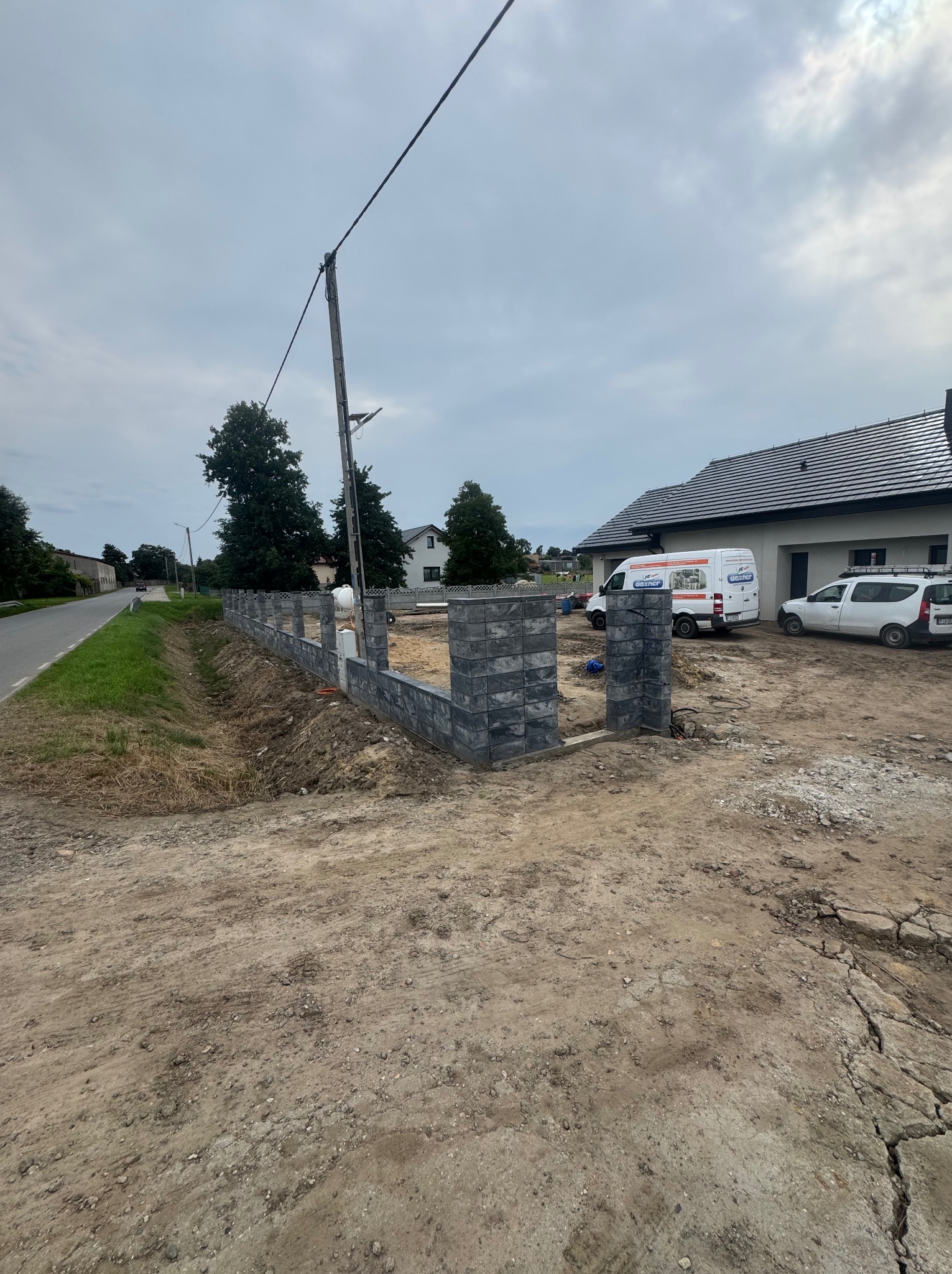 Wznoszona, szara, segmentowa konstrukcja ogrodzenia na posesji. W tle dom z szarą dachówką, dwa białe samochody dostawcze i słup energetyczny.