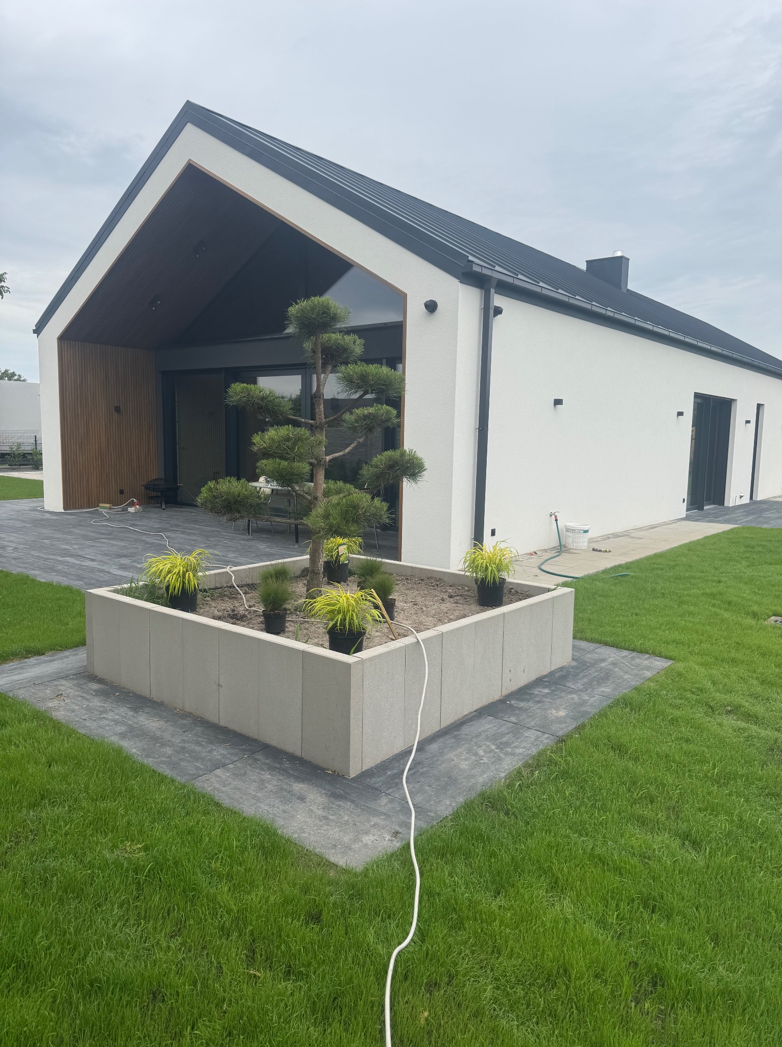 Nowoczesny dom z białymi ścianami i czarnym dachem, z dużymi oknami i drewnianą elewacją, otoczony trawnikiem i ozdobnym klombem z roślinami.