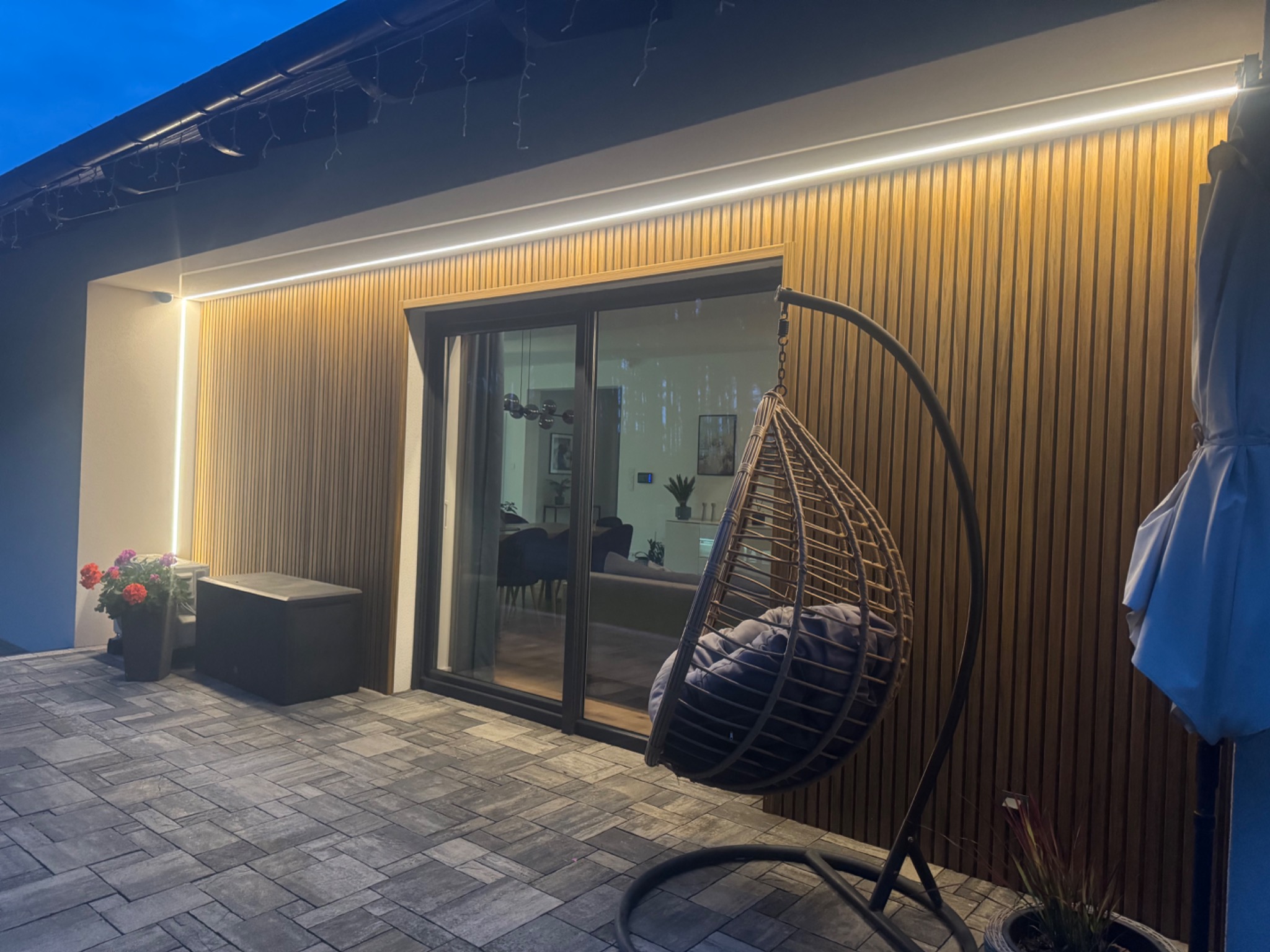 Nowoczesna elewacja domu z drewnianymi panelami, oświetleniem LED i szarą kostką brukową. Widoczny fragment tarasu z wiszącym fotelem i kwiatami.