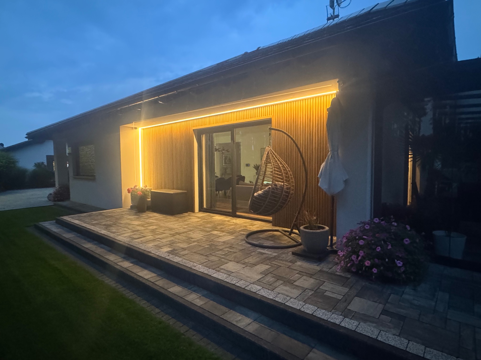 Nowoczesny dom z podświetloną elewacją drewnianą i tarasem z kostki brukowej. W tle widoczny ogród i wiszące krzesło.