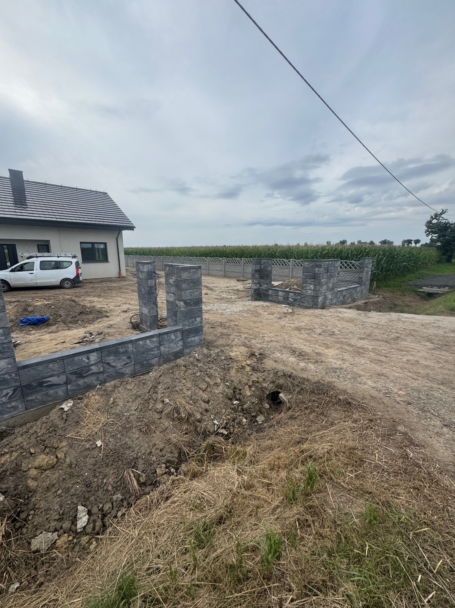 Budowa ogrodzenia z szarych bloczków betonowych na tle domu i pola. Widoczne słupki i fragmenty muru, w tle zaparkowany biały samochód.
