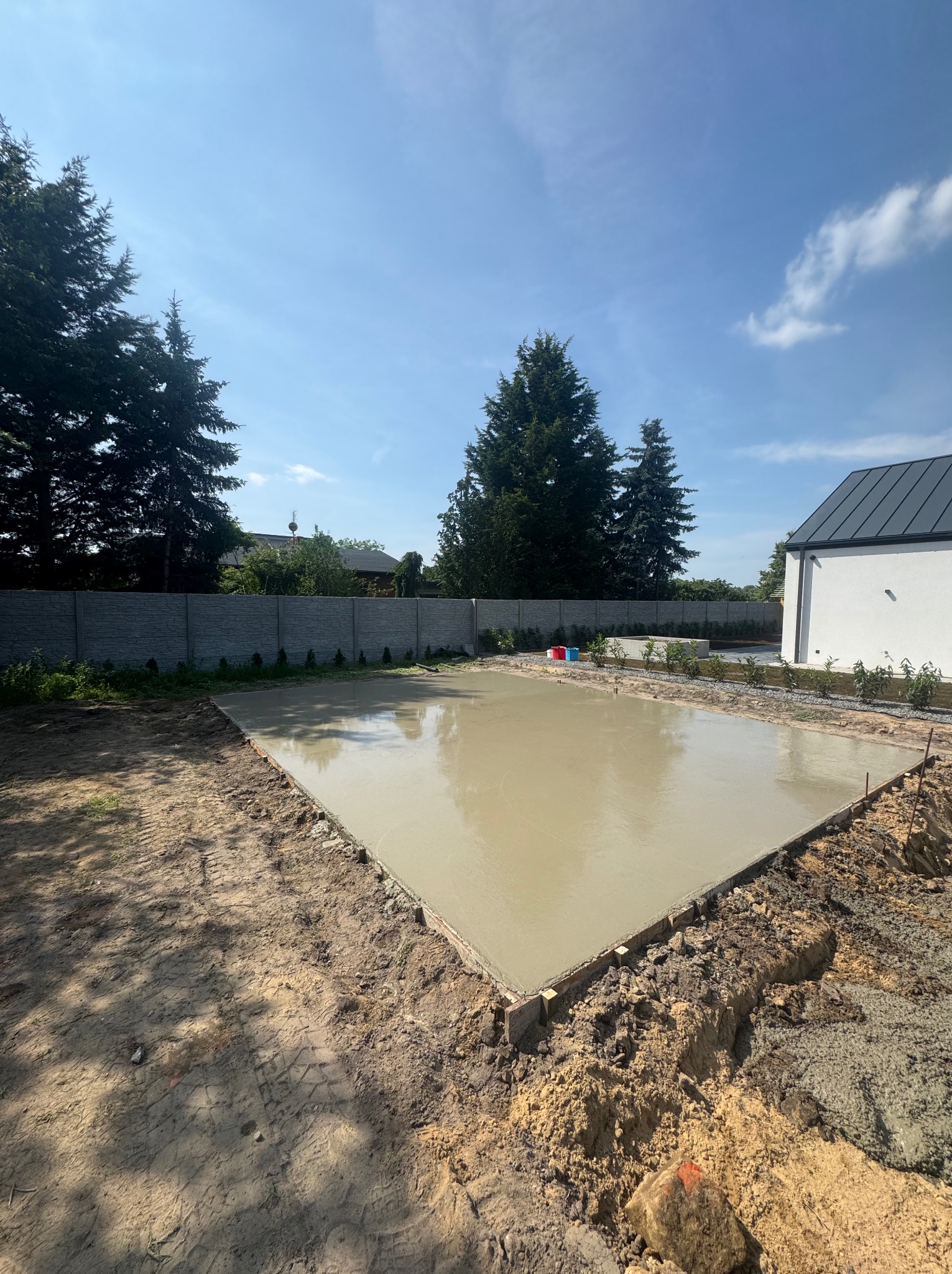 Świeżo wylana, gładka płyta fundamentowa pod budowę domu, otoczona ziemią i drewnianym szalunkiem, w tle widoczne drzewa i fragment budynku.