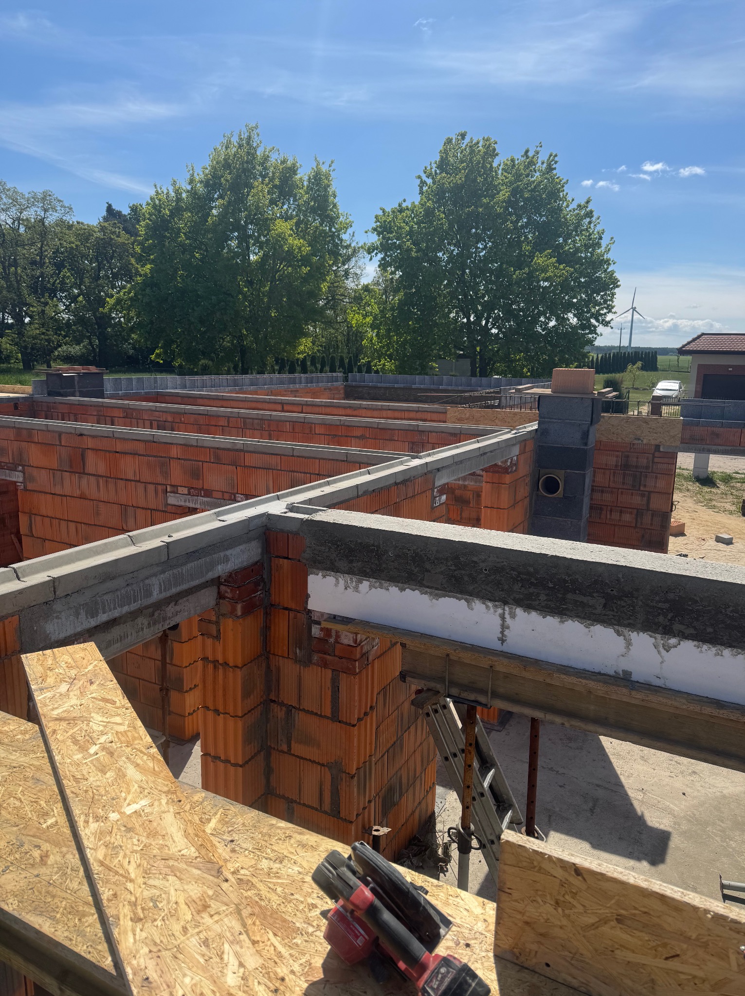 Wznoszenie ścian z czerwonej cegły na budowie domu jednorodzinnego. Widoczne szalunki, narzędzia i elementy konstrukcyjne. W tle drzewa i turbina wiatrowa.