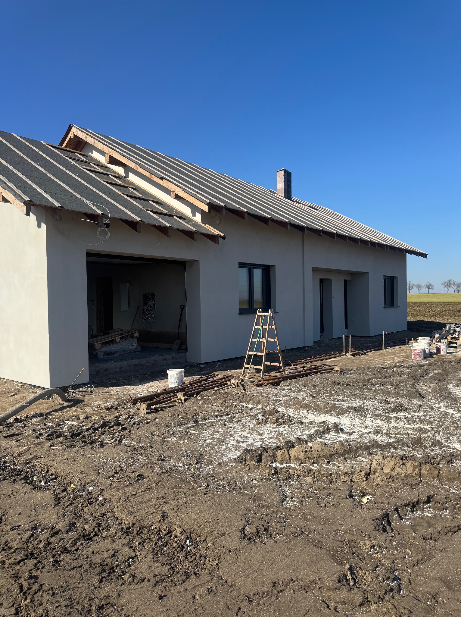 Surowy, otynkowany dom jednorodzinny w trakcie budowy, z widocznym dachem w budowie, oknami i drabiną opartą o elewację. W tle pola i błękitne niebo.