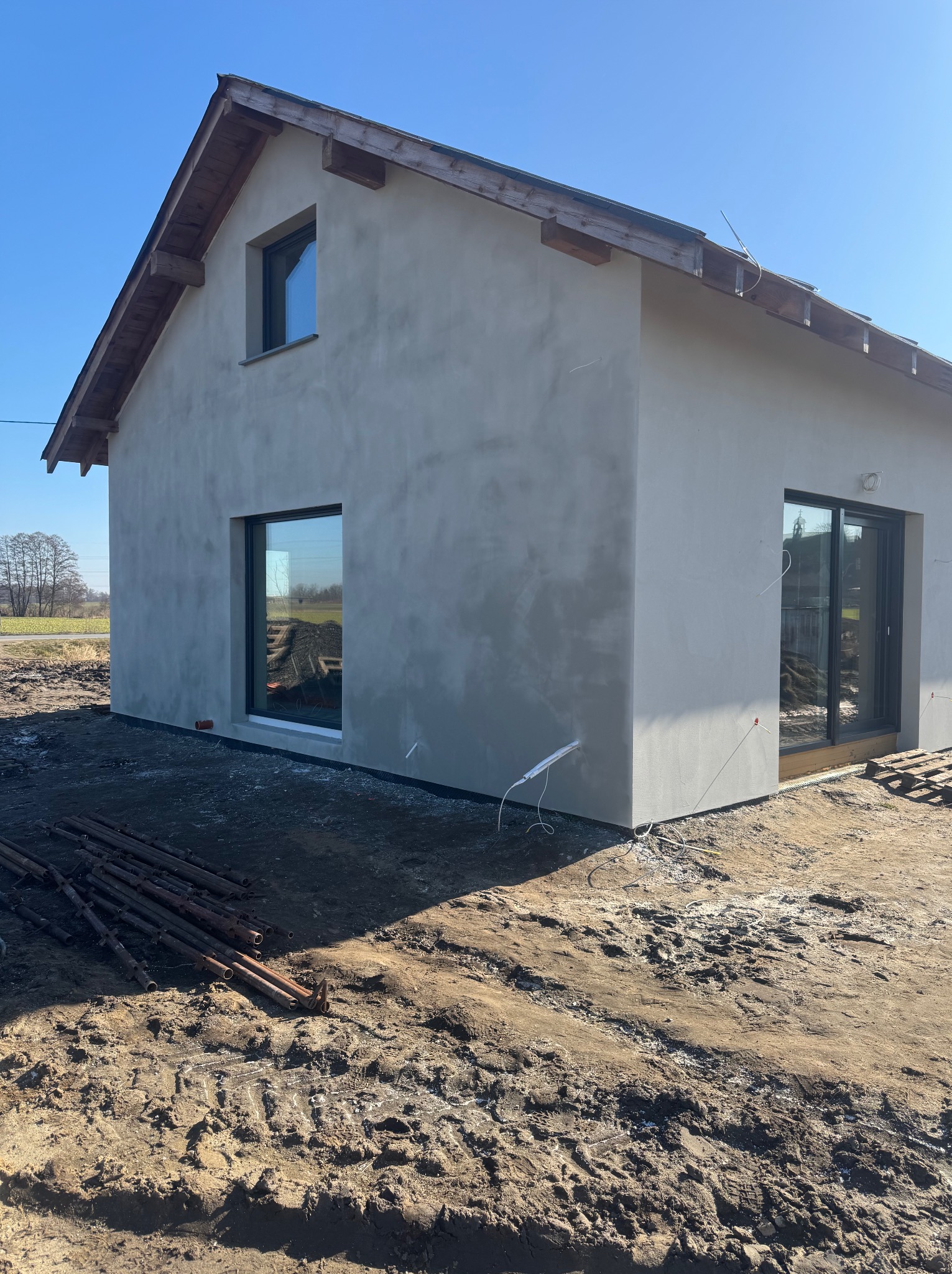 Surowy, otynkowany dom jednorodzinny z drewnianą konstrukcją dachu, duże okna, teren wokół domu w trakcie prac ziemnych. Słoneczny dzień, błękitne niebo.