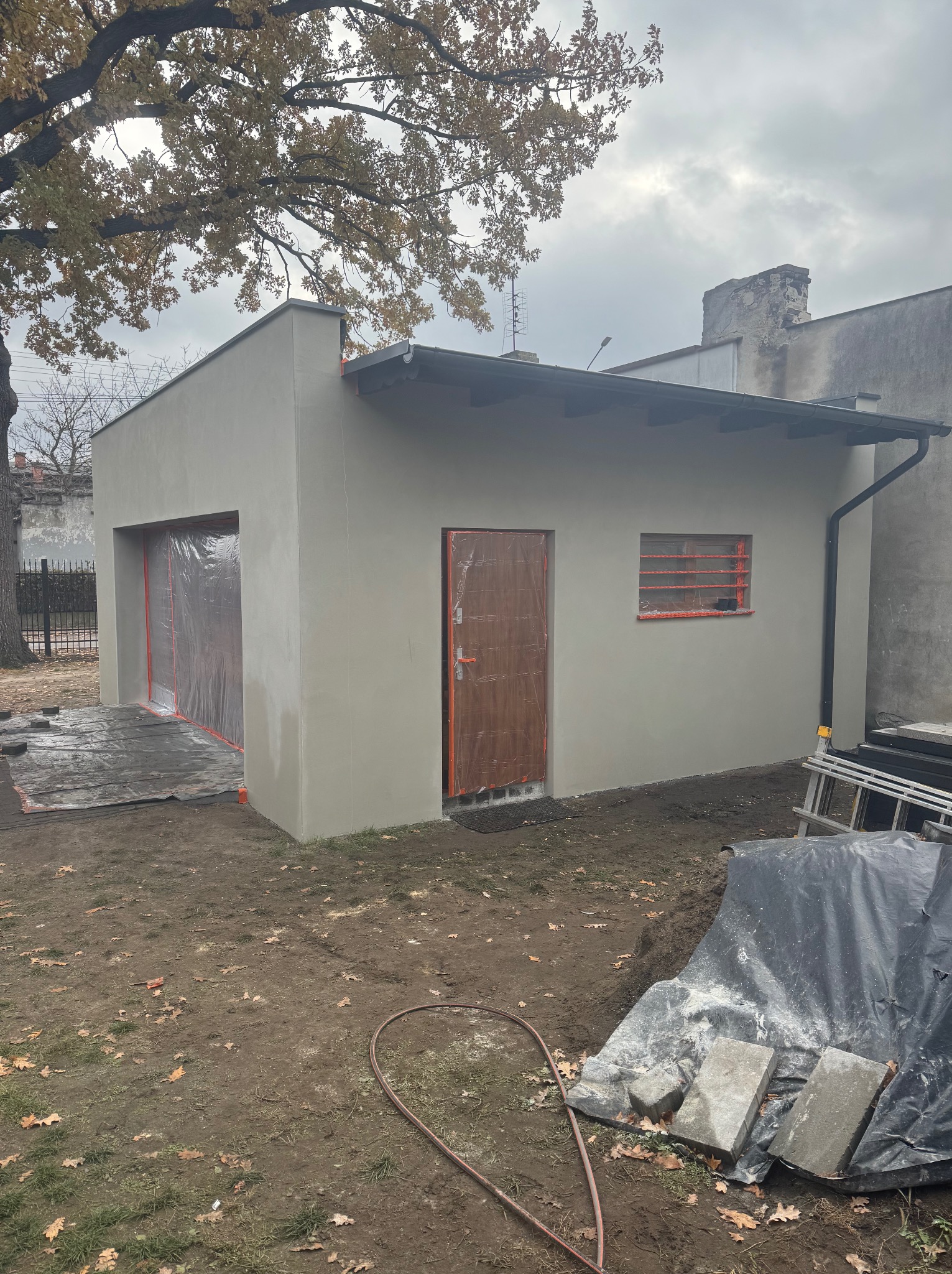 Otynkowany budynek gospodarczy zadaszony, z nową stolarką okienną i drzwiową zabezpieczoną folią, obok drabina i materiały budowlane.