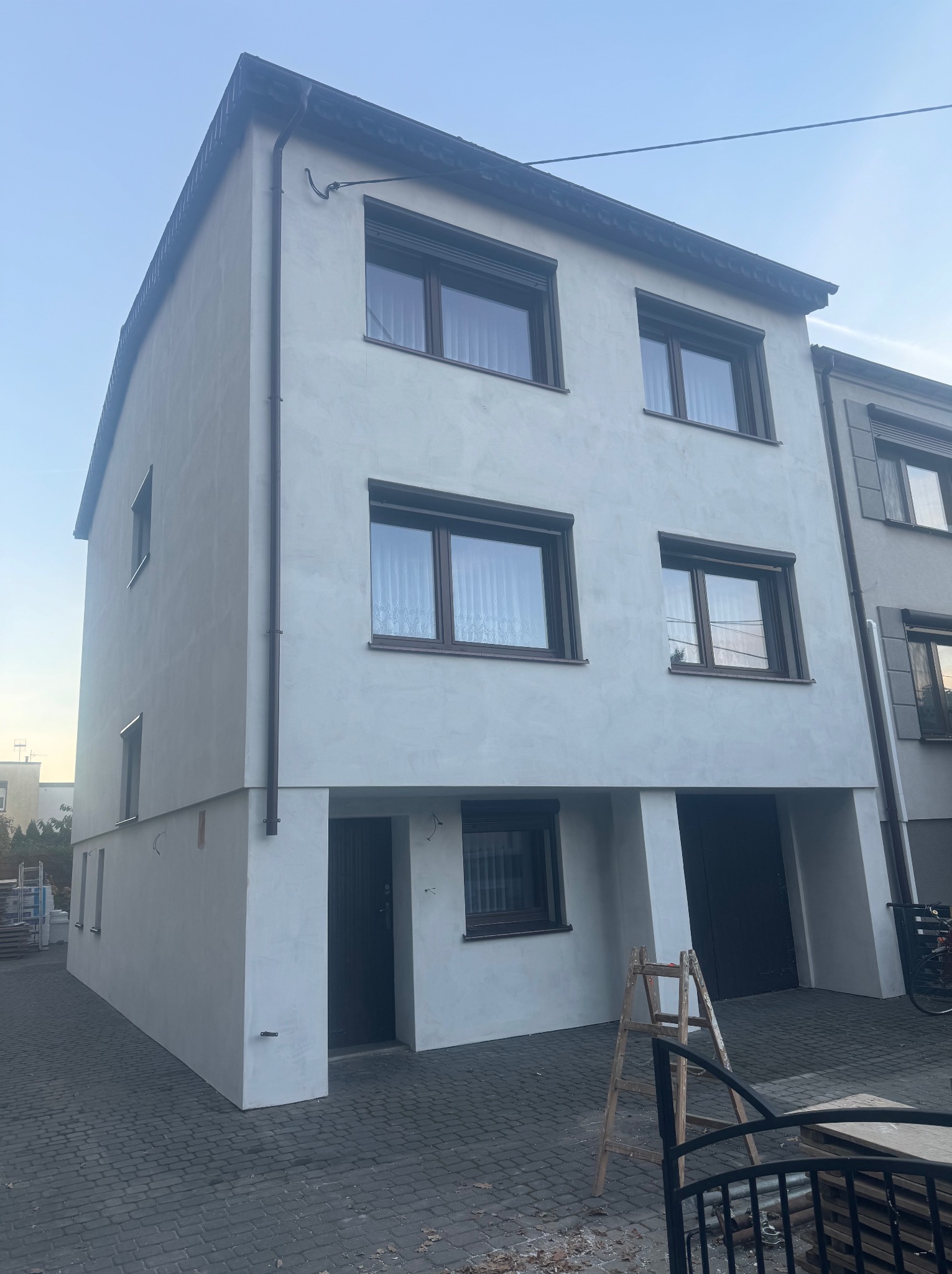 Ocieplony, dwukondygnacyjny dom z nową, jasną elewacją i brązowymi oknami. Na podjeździe stoi drewniana drabina. Widok z dołu, w tle niebieskie niebo.