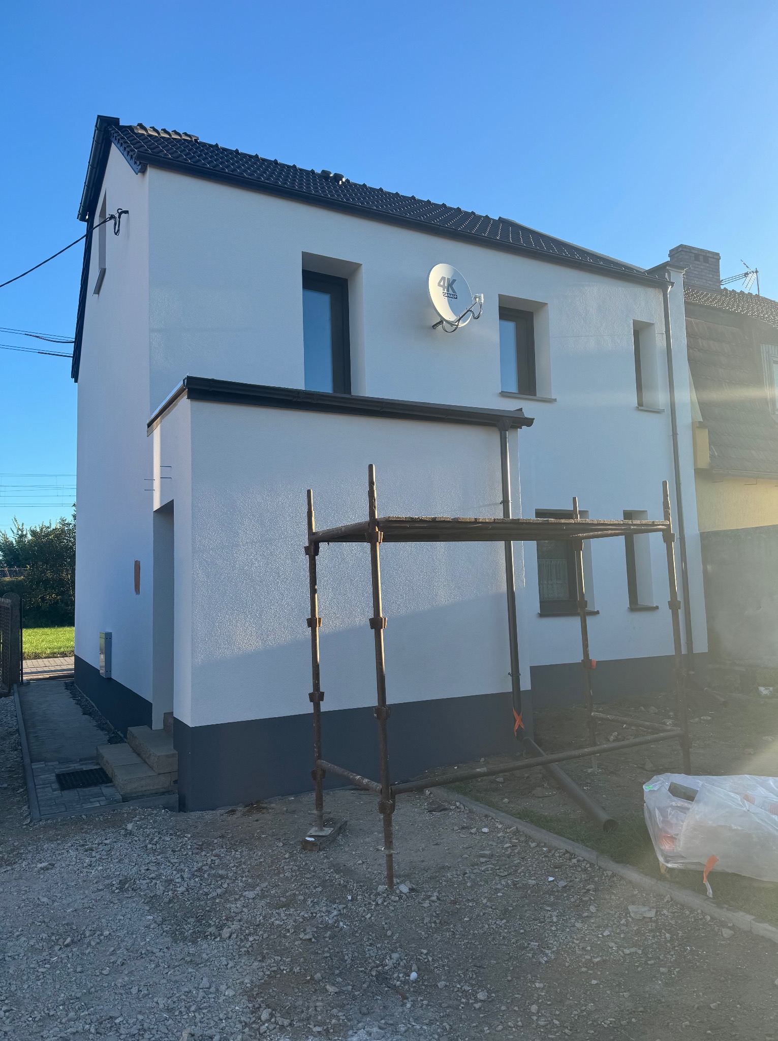 Dwukondygnacyjny dom z białą elewacją i ciemną podmurówką, widoczny z rusztowaniem. Na ścianie antena satelitarna. Elewacja po malowaniu w słoneczny dzień.